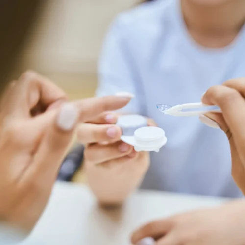 Close up of female oculist hand holding tweezers with contact lens while little boy with container sitting at the table